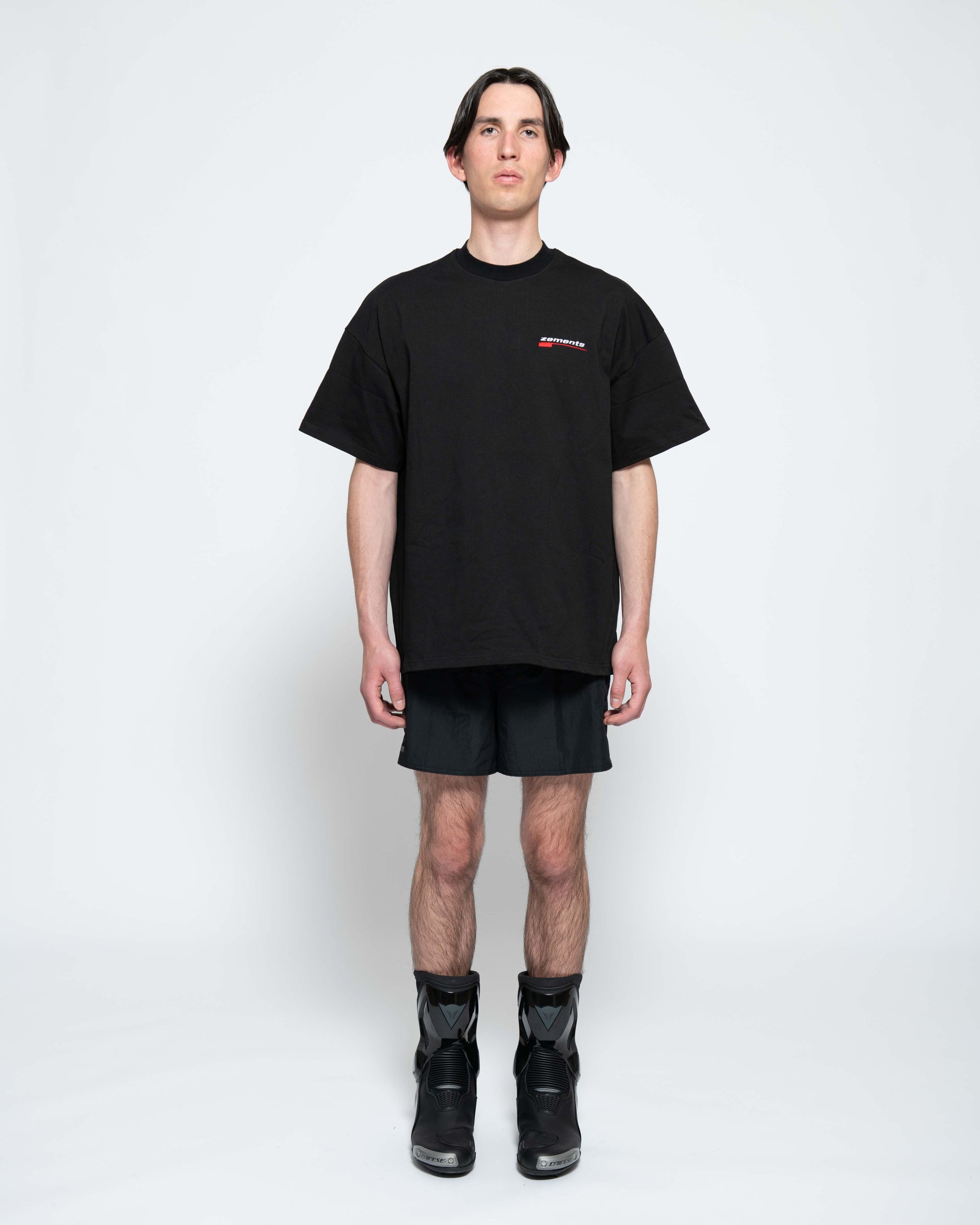 Frontalansicht, Ganzkörperbild. Männliches Model, mit einem schwarzen oversized "Swoosh Zements" T-shirt. Oben Links auf Brust-Höhe, ist das "Swoosh Zements-Logo" in Rot/Weiß klein aufgestickt. Dazu trägt das Model eine kurze schwarze Short und schwarze Bikerstiefel