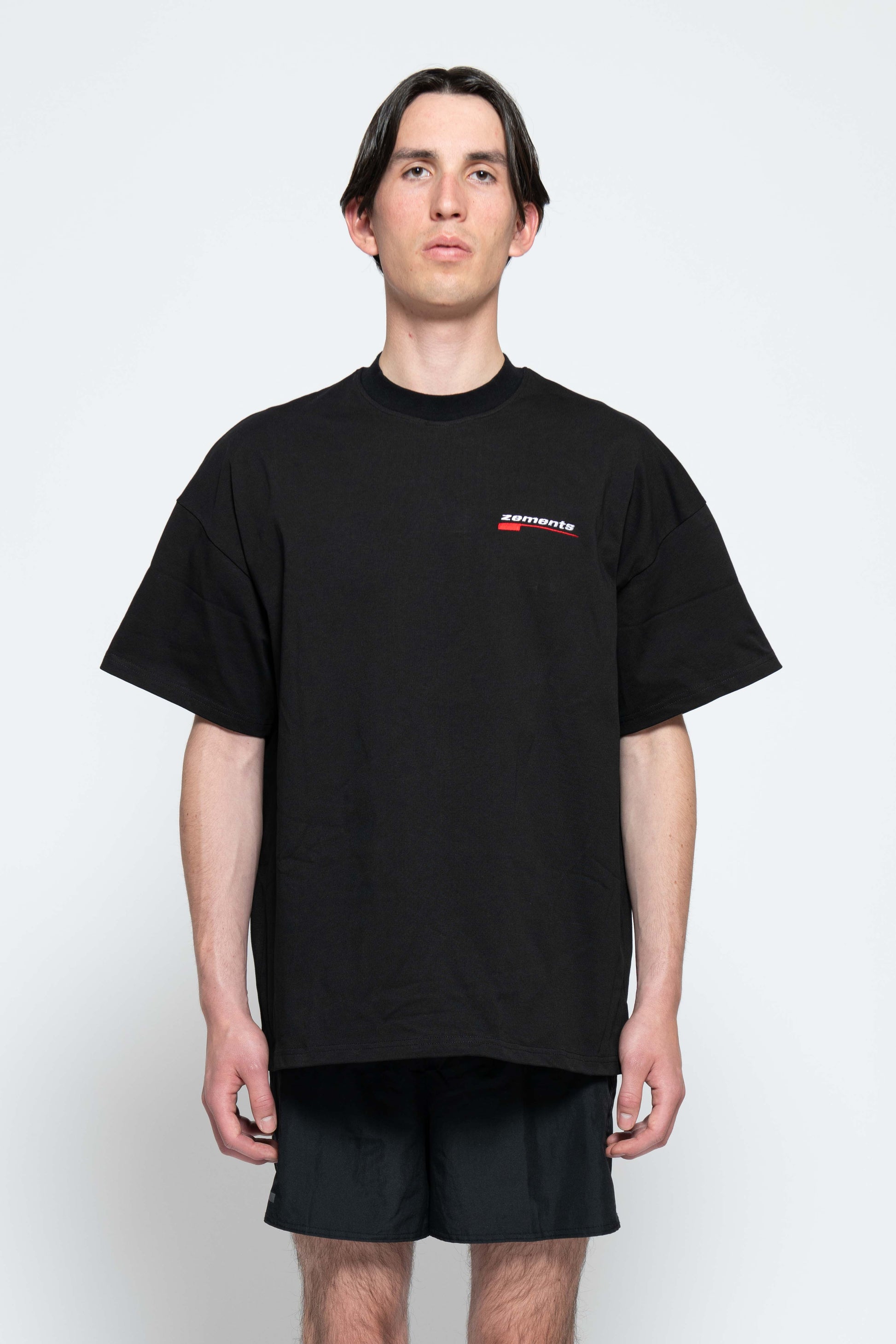 Frontalansicht, Halbkörperbild. Männliches Model, mit einem schwarzen oversized "Swoosh Zements" T-shirt. Oben Links auf Brust-Höhe, ist das "Swoosh Zements-Logo" in Rot/Weiß klein aufgestickt.