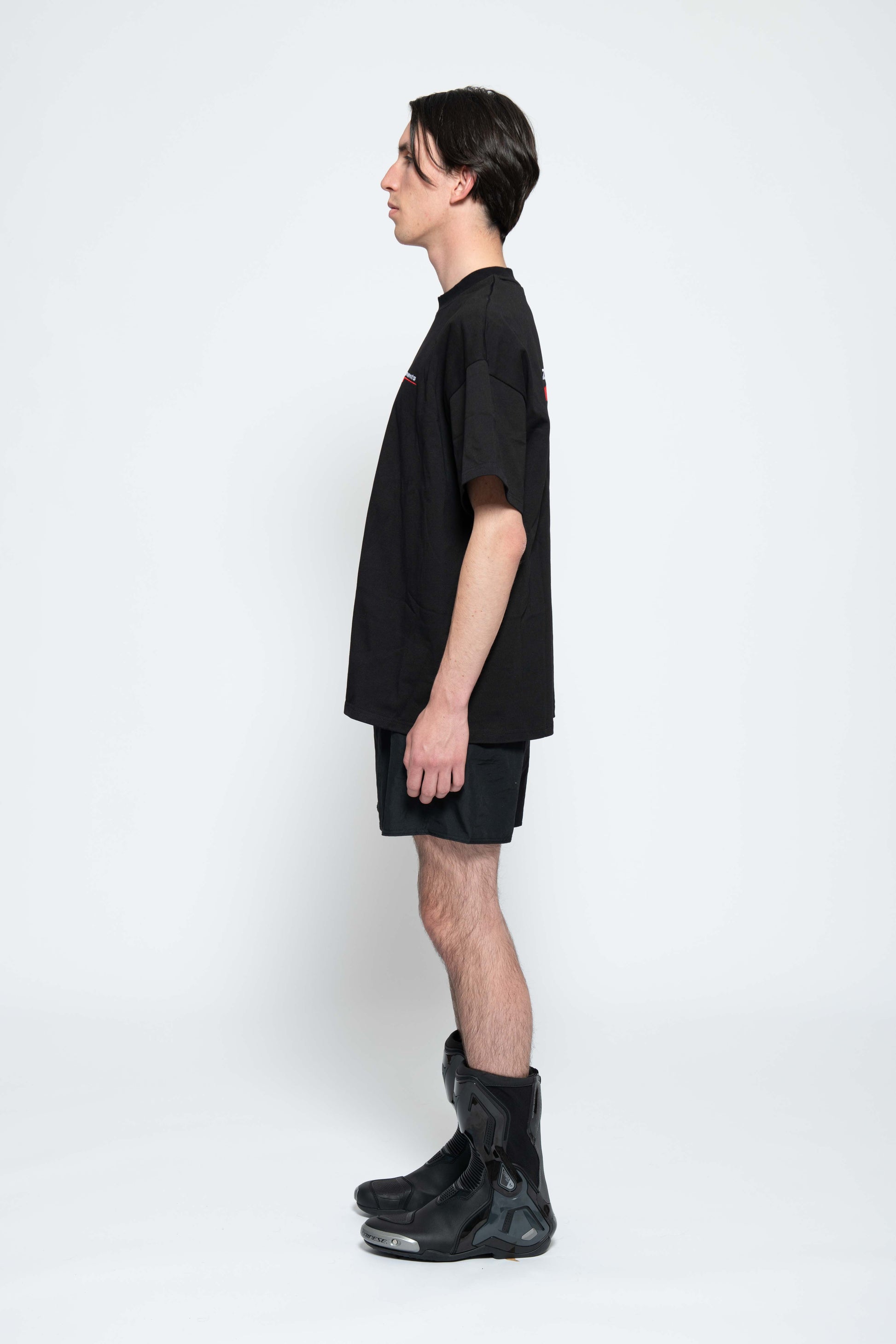 Seitenansicht, Ganzkörperbild. Männliches Model, mit einem schwarzen oversized "Swoosh Zements"T-shirt.  Dazu trägt das Model eine kurze schwarze Short und schwarze Bikerstiefel