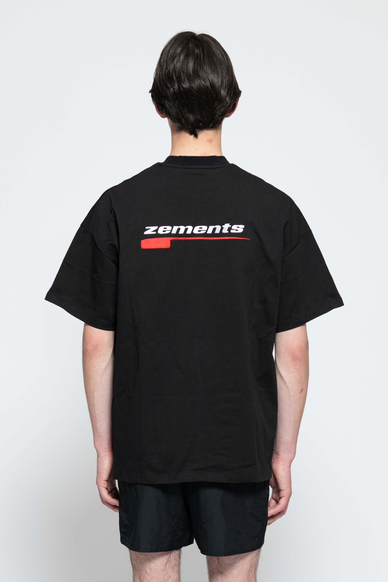 Frontalansicht, Ganzkörperbild. Männliches Model, mit einem schwarzen oversized "Swoosh Zements" T-shirt. Oben Links auf Brust-Höhe, ist das "Swoosh Zements-Logo" in Rot/Weiß klein aufgestickt. Dazu trägt das Model eine kurze schwarze Short und schwarze Bikerstiefel