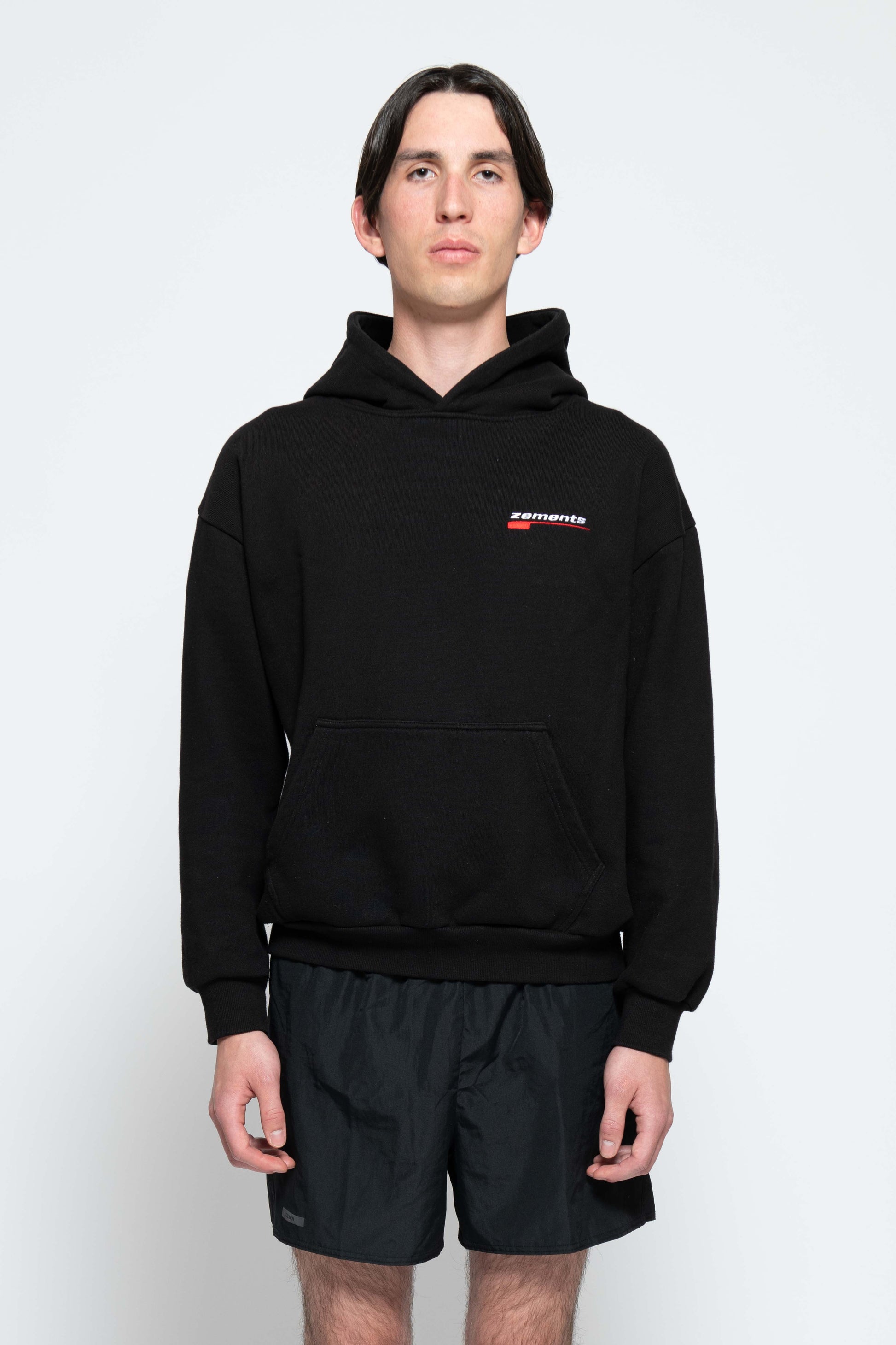 Frontalansicht, Halbkörperbild. Männliches Model, mit einem schwarzen Unisex "Swoosh Zements" Hoodie. Oben Links auf Brust-Höhe, ist das "Swoosh Zements-Logo" in Rot/Weiß klein aufgestickt.