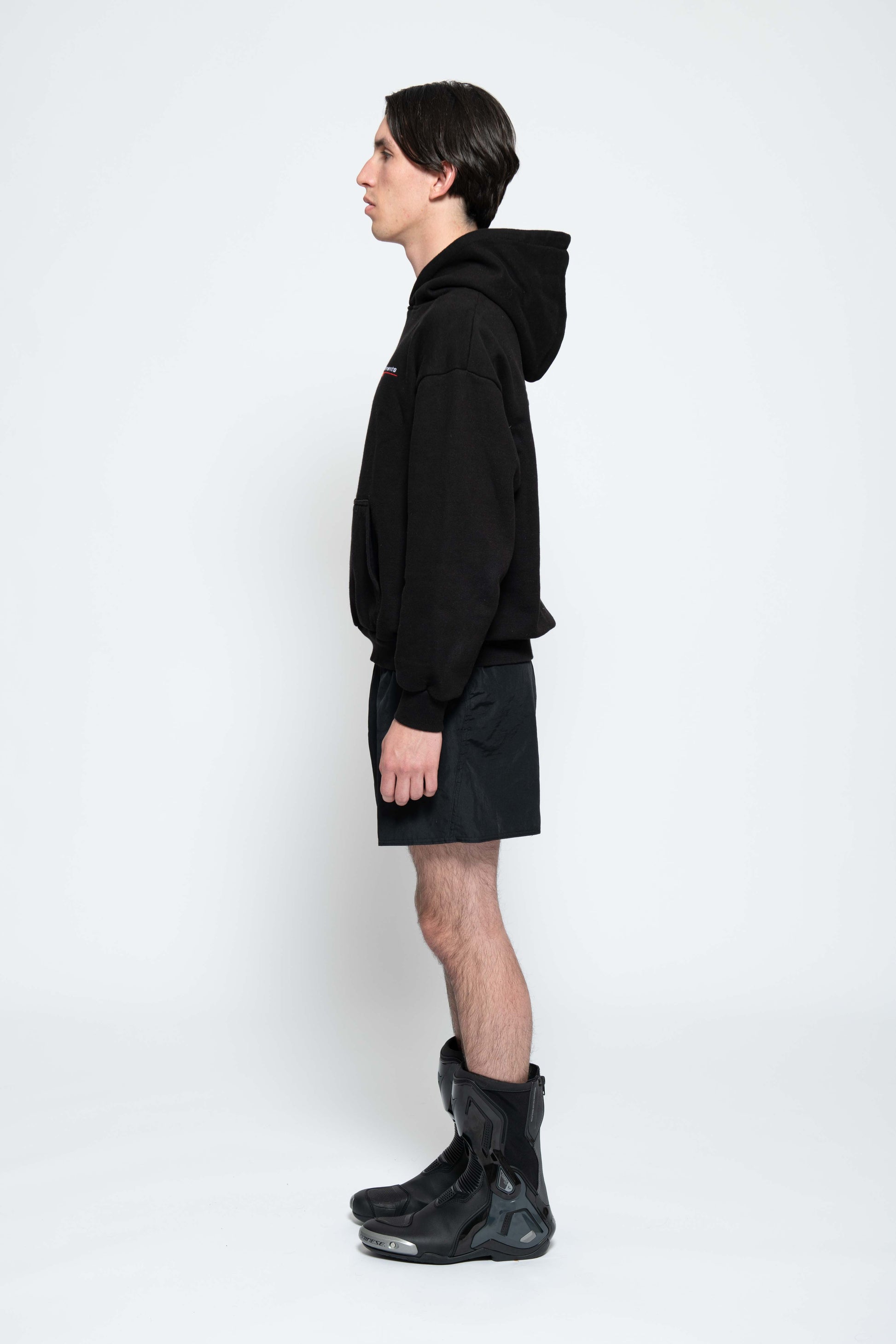 Seitenansicht, Ganzkörperbild. Männliches Model, mit einem schwarzen Unisex "Swoosh Zements" Hoodie.  Dazu trägt das Model eine kurze schwarze Short und schwarze Bikerstiefel