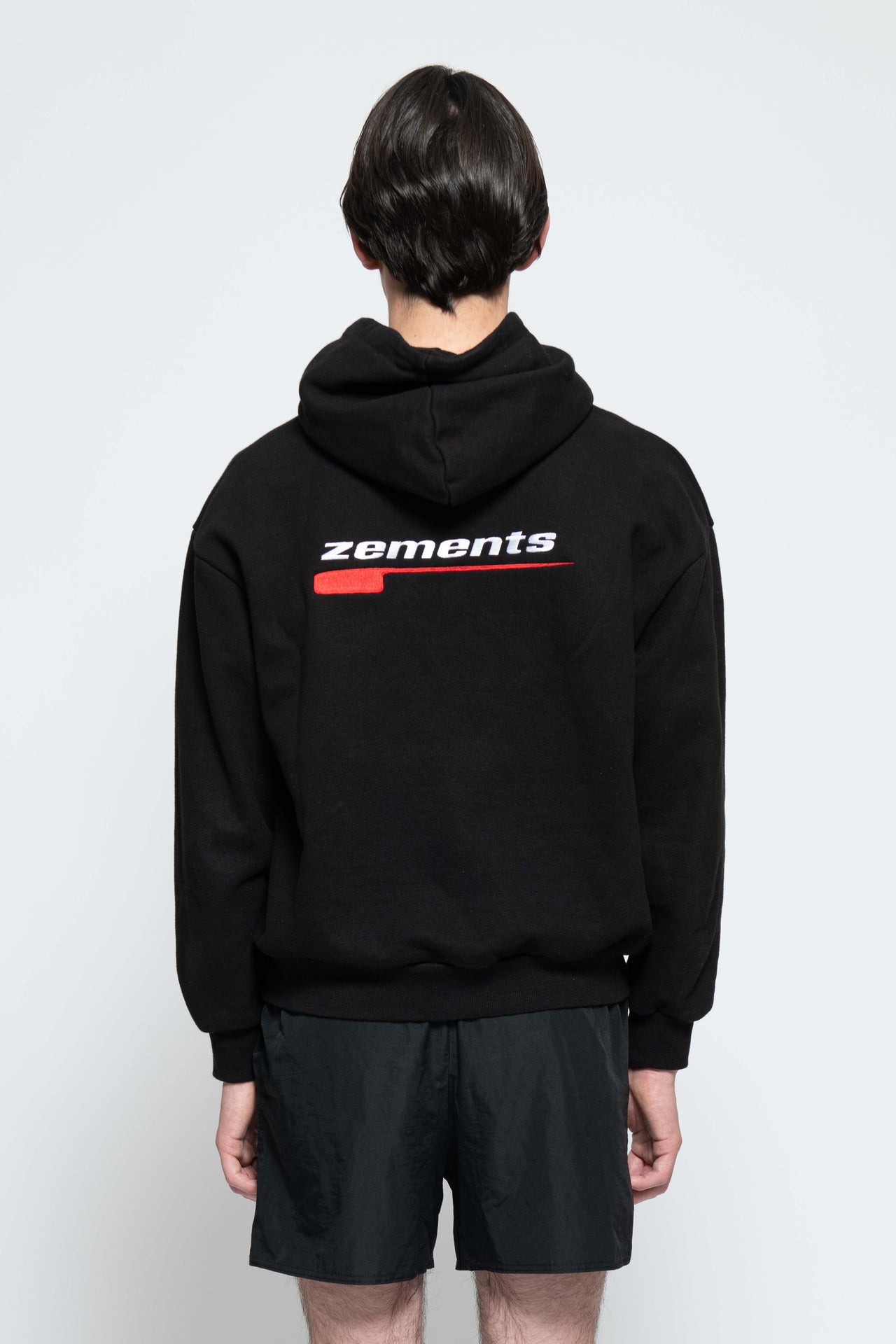 Frontalansicht, Ganzkörperbild. Männliches Model, mit einem schwarzen Unisex "Swoosh Zements" Hoodie. Oben Links auf Brust-Höhe, ist das "Swoosh Zements-Logo" in Rot/Weiß klein aufgestickt. Dazu trägt das Model eine kurze schwarze Short und schwarze Bikerstiefel