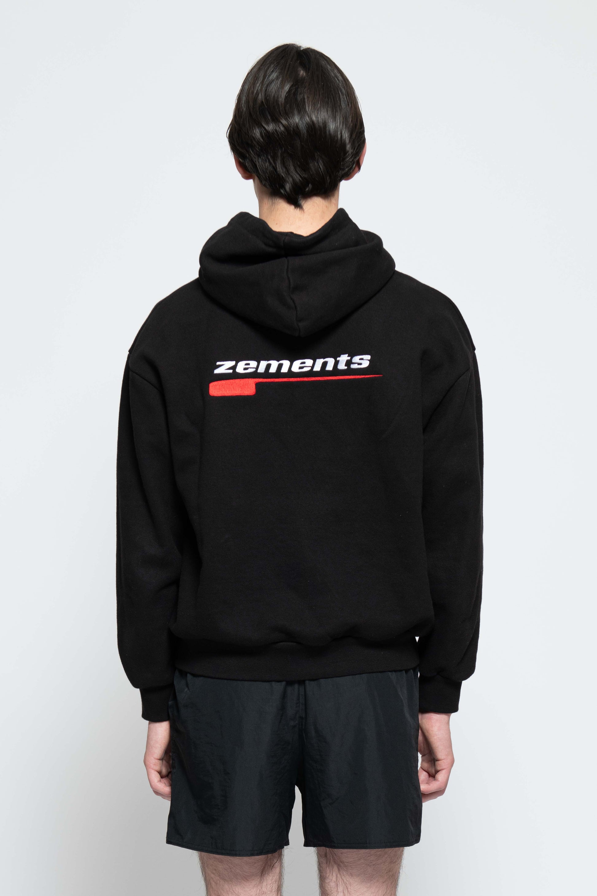 Rückenansicht, Halbkörperbild. Männliches Model, mit einem schwarzen Unisex "Swoosh Zements" Hoodie. In der Mitte auf Schulterblatt-Höhe, ist das "Swoosh Zements-Logo" in Rot/Weiß groß aufgestickt.