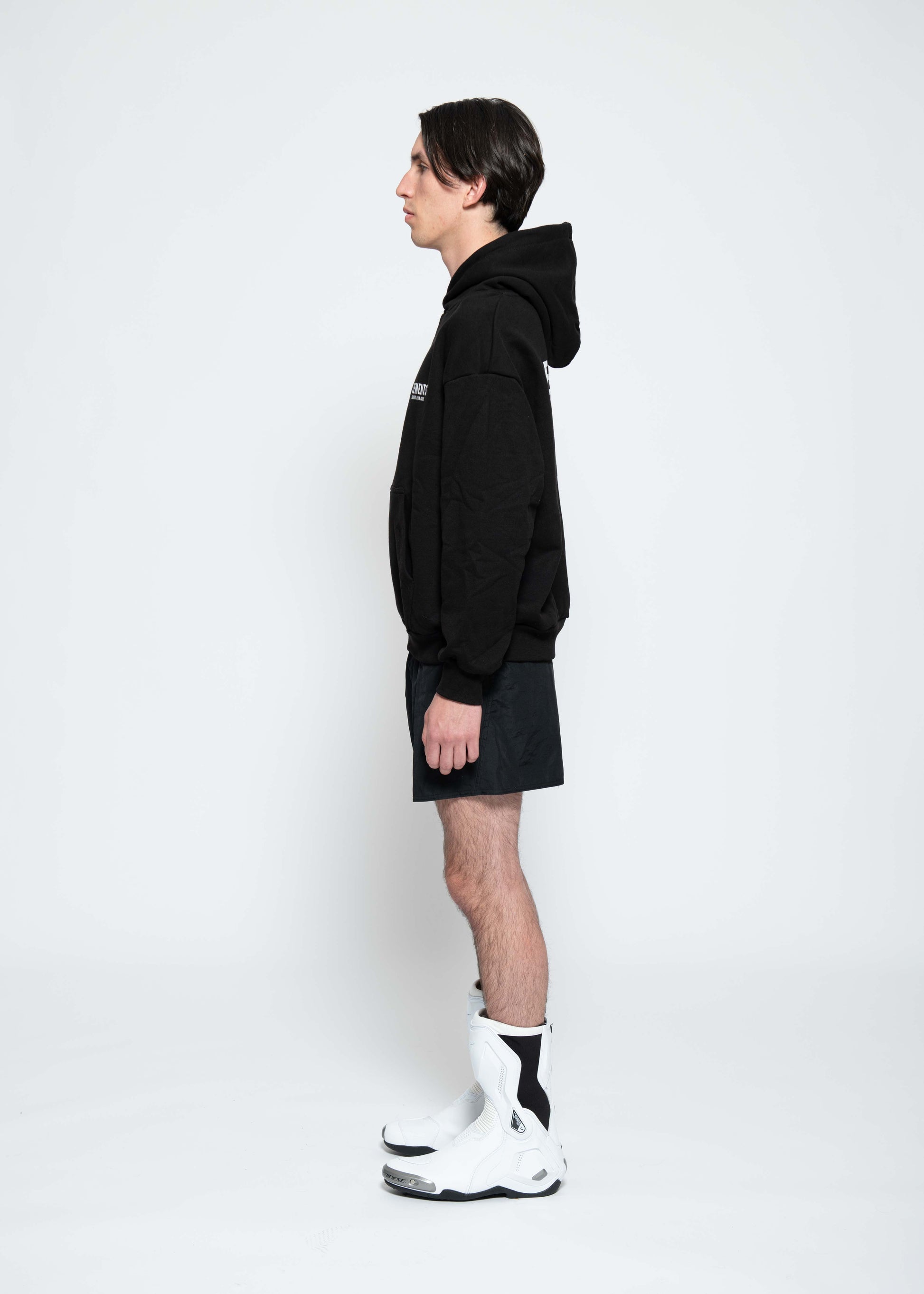 Frontalansicht, Ganzkörperbild. Männliches Model, mit einem schwarzen unisex „Sweet Pain Issue“ Hoodie. Dazu trägt das Model eine kurze schwarze Short und schwarze Bikerstiefel