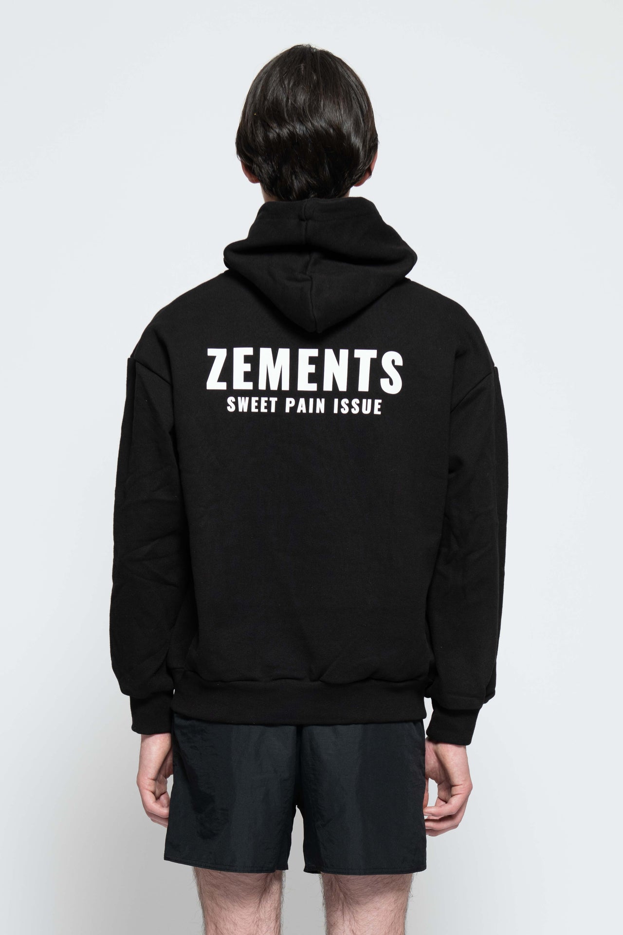 Frontalansicht, Ganzkörperbild. Männliches Model, mit einem schwarzen unisex „Sweet Pain Issue“ Hoodie. Oben Links auf Brust-Höhe, ist das „Zements-Logo„ und drunter das „Sweet Pain Issue-Logo“ in Weiß klein aufgedruckt. Dazu trägt das Model eine kurze schwarze Short und schwarze Bikerstiefel