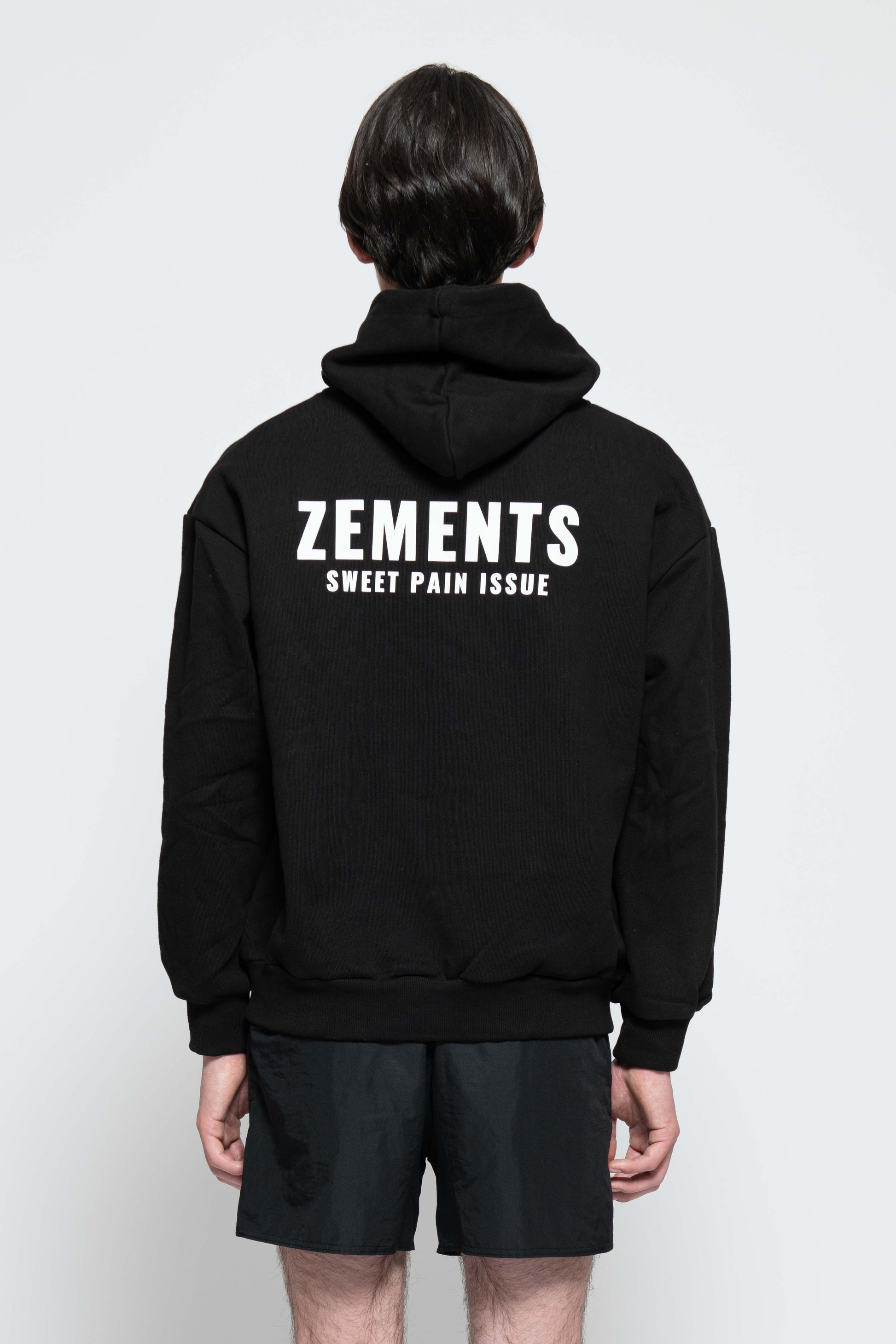 Rückenansicht, Halbkörperbild. Männliches Model, mit einem schwarzen Unisex „Sweet Pain Issue“ Hoodie. In der Mitte auf Schulterblatt-Höhe, ist das „Zements-Logo„ und drunter das „Sweet Pain Issue-Logo“ in Weiß groß aufgedruckt.