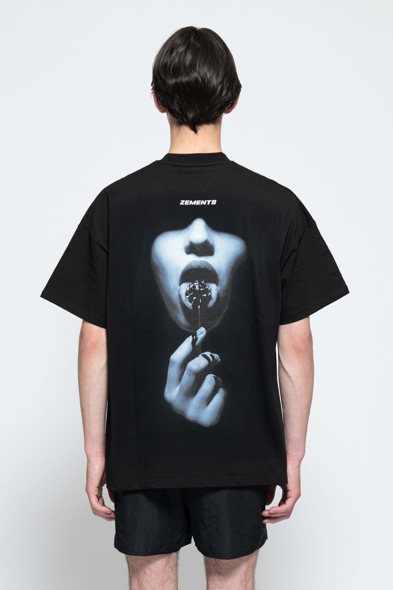 Frontalansicht, Ganzkörperbild. Männliches Model, mit einem schwarzen oversized „Sweet Pain Issue“ T-shirt. Oben Links auf Brust-Höhe, ist das „Zements-Logo" in Weiß klein aufgedruckt. Dazu trägt das Model eine kurze schwarze Short und schwarze Bikerstiefel