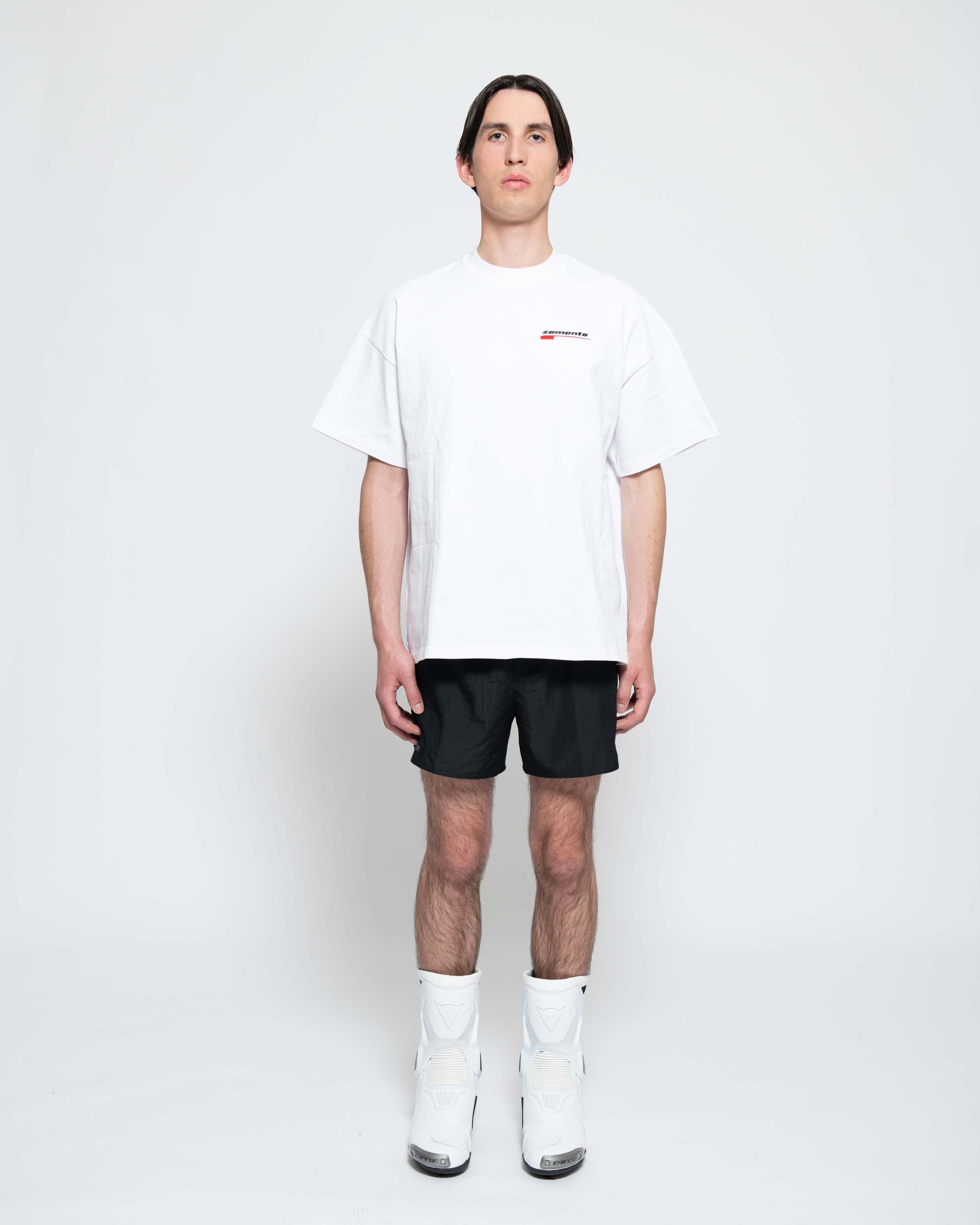 Frontalansicht, Ganzkörperbild. Männliches Model, mit einem weißen oversized "Swoosh Zements" T-shirt. Oben Links auf Brust-Höhe, ist das "Swoosh Zements-Logo" in Rot/Schwarz klein aufgestickt. Dazu trägt das Model eine kurze schwarze Short und schwarze Bikerstiefel