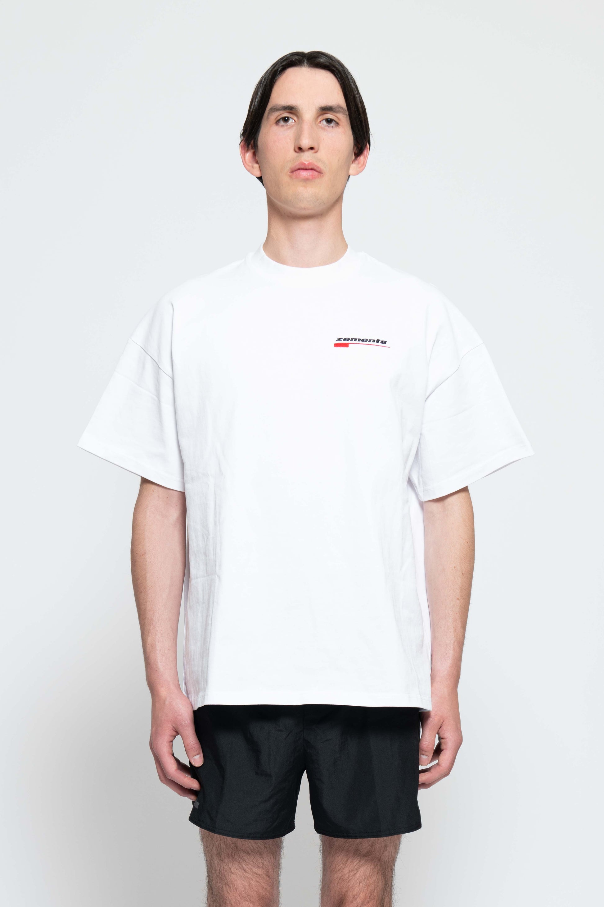 Frontalansicht, Halbkörperbild. Männliches Model, mit einem weißen oversized "Swoosh Zements" T-shirt. Oben Links auf Brust-Höhe, ist das "Swoosh Zements-Logo" in Rot/Schwarz klein aufgestickt.