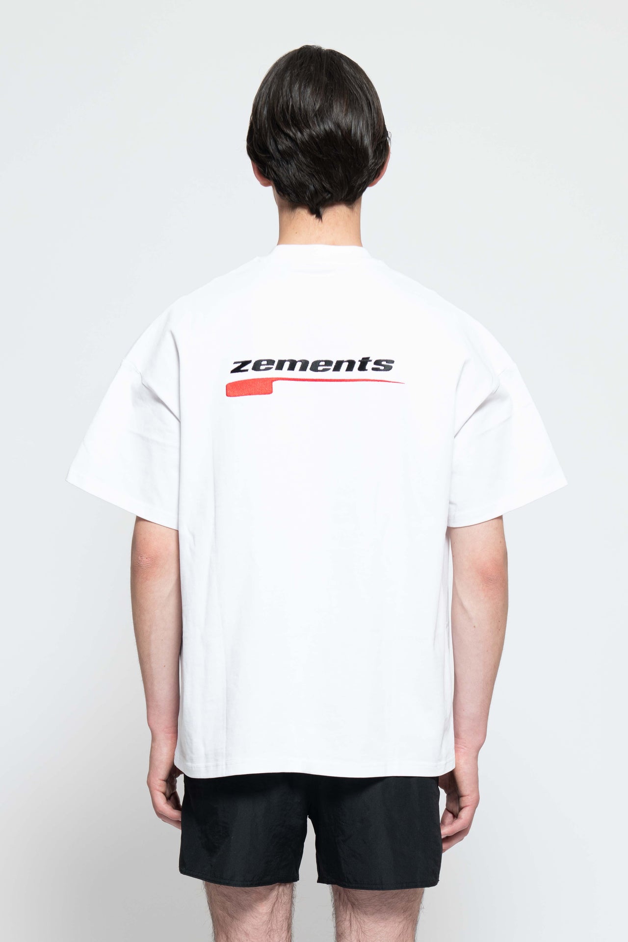 Frontalansicht, Ganzkörperbild. Männliches Model, mit einem weißen oversized "Swoosh Zements" T-shirt. Oben Links auf Brust-Höhe, ist das "Swoosh Zements-Logo" in Rot/Schwarz klein aufgestickt. Dazu trägt das Model eine kurze schwarze Short und schwarze Bikerstiefel