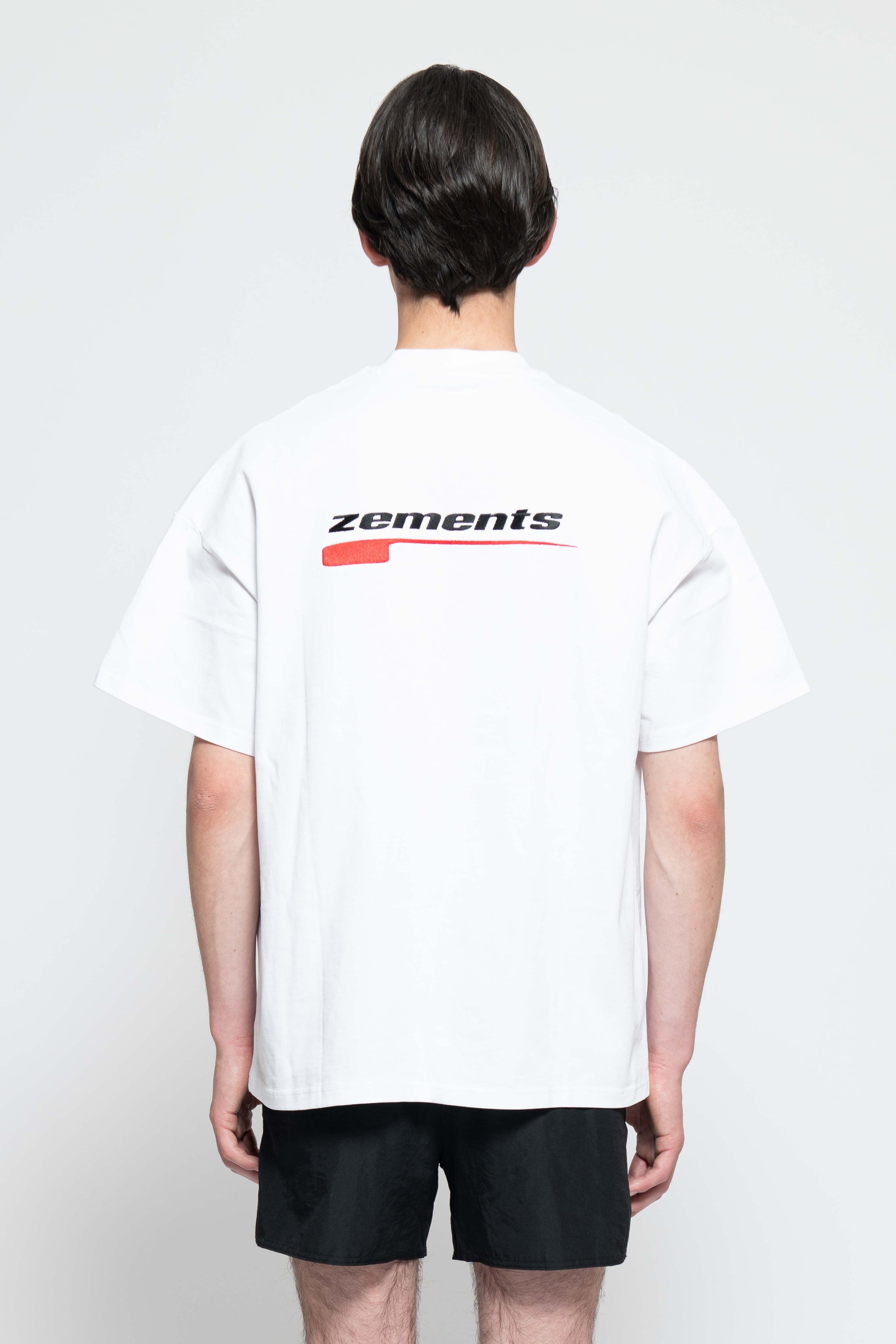 Rückenansicht, Halbkörperbild. Männliches Model, mit einem weißen oversized "Swoosh Zements" T-shirt. In der Mitte auf Schulterblatt-Höhe, ist das "Swoosh Zements-Logo" in Rot/Schwarz groß aufgestickt.